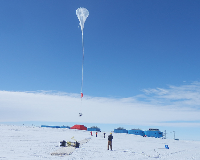 Balloon launch at remote Antarctica base against a clear blue sky, with scientists observing equipment on the snow.