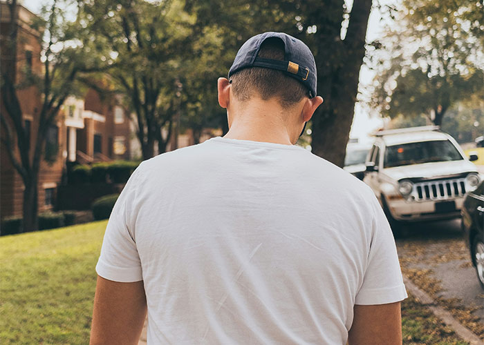 Back view of a person walking on a sidewalk, wearing a cap and white shirt, conveying a sense of needing to leave quickly.