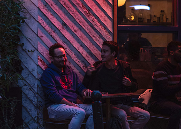 Two men sitting outside a café under red light, sharing a conversation about scary experiences.