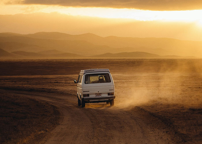 A van driving on a dusty road through a vast desert landscape at sunset.