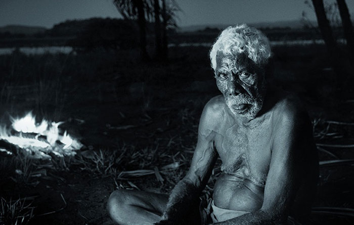 Elderly man sitting by a campfire at night, conveying a sense of disturbing facts and contemplation.