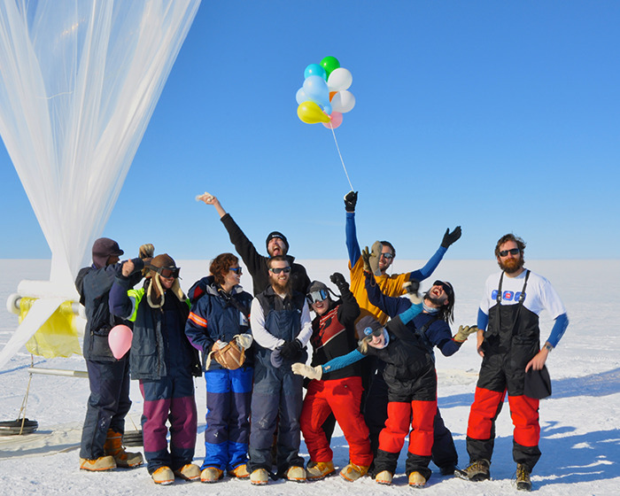 Scientists in Antarctica pose cheerfully with balloons and winter gear against a snowy landscape.