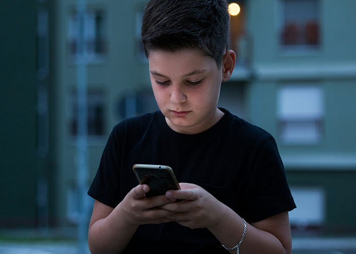 Young boy focused on his phone, wearing a black t-shirt, standing outdoors with buildings in the background.