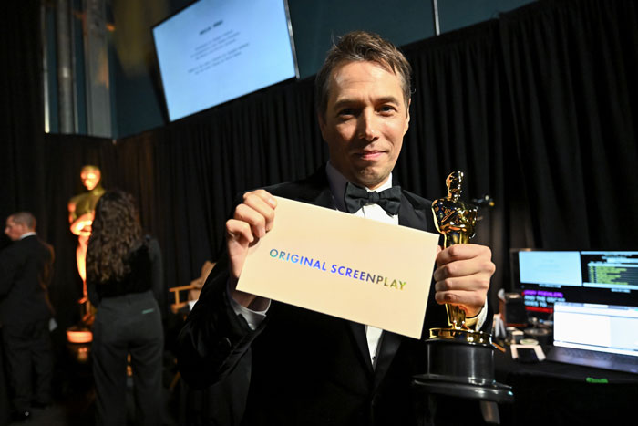 Winner holding Oscar 2025 for Original Screenplay, smiling backstage with award envelope.