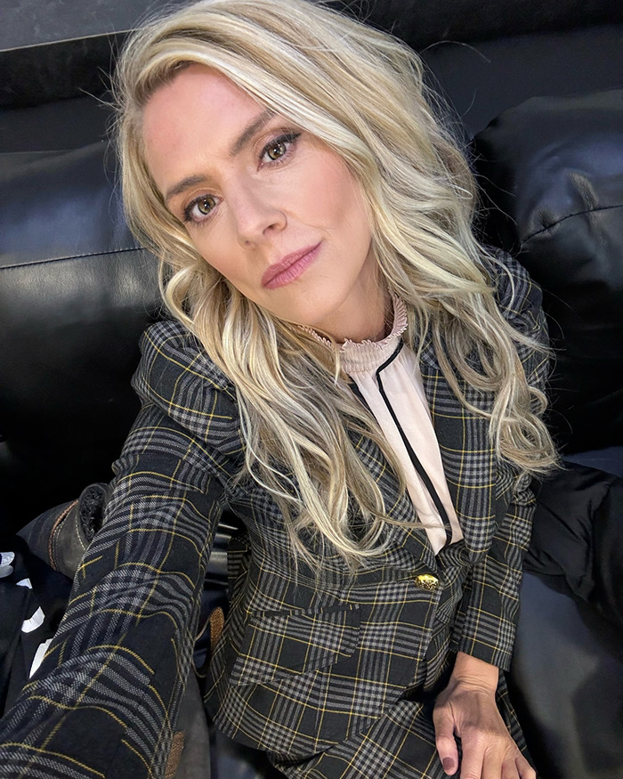 Blonde woman in checkered blazer, seated on a black couch, poses for selfie.