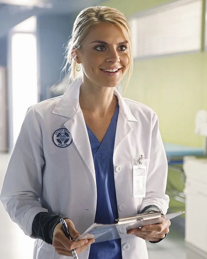 Woman in a white lab coat holding a clipboard, smiling in a medical setting.