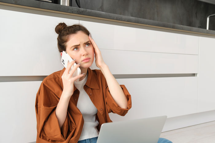 Woman sitting on kitchen floor, worried, holding phone to ear with laptop nearby, discussing friend's whereabouts.