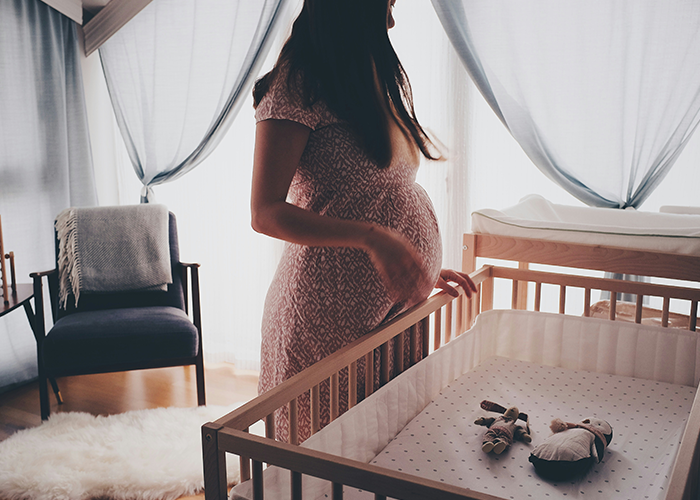 Pregnant woman in a softly lit nursery, standing by a wooden crib, with toys inside, wearing a patterned dress.