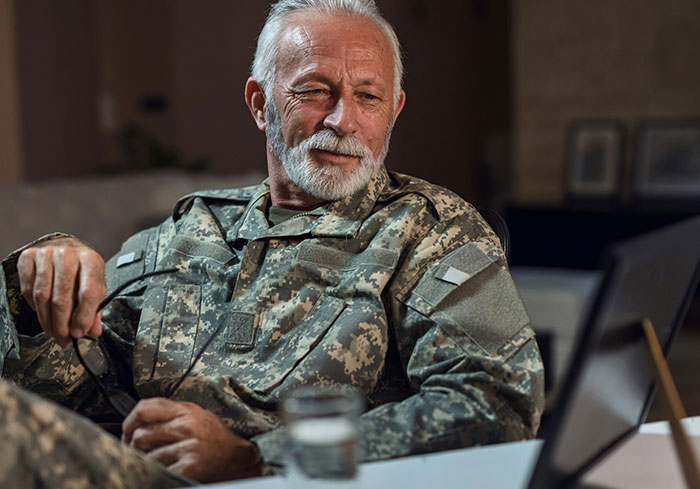 Elderly man in military uniform sitting at a desk with a laptop, holding glasses, depicting stories about bosses from hell.