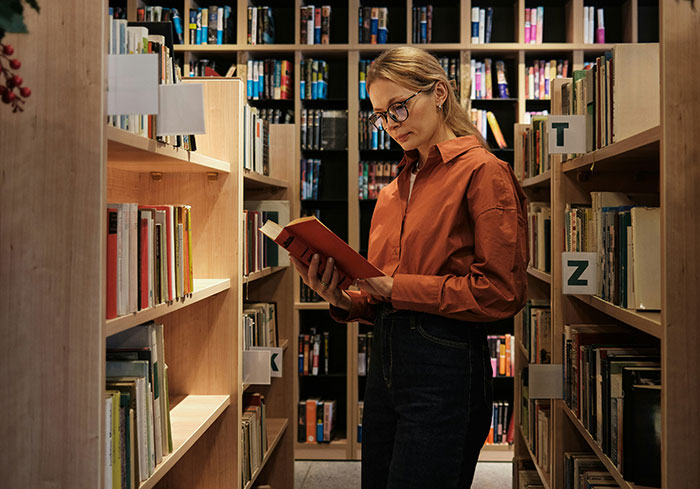 A woman in glasses and an orange shirt reads a book in a library aisle.