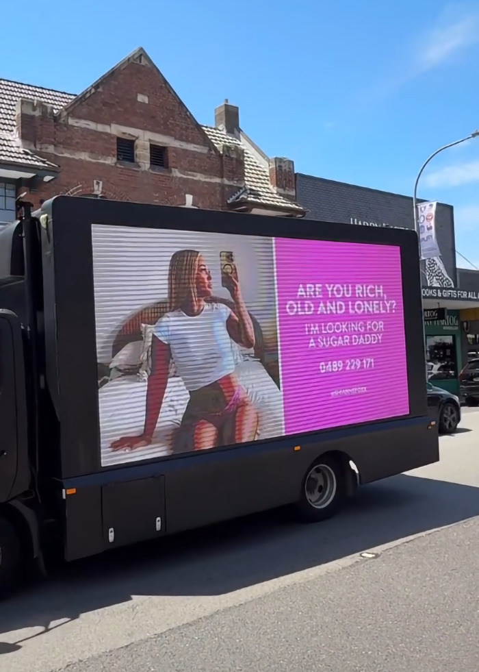 Mobile billboard featuring "Bikini Tradie" ad seeking a sugar daddy, displayed on a truck in urban setting.