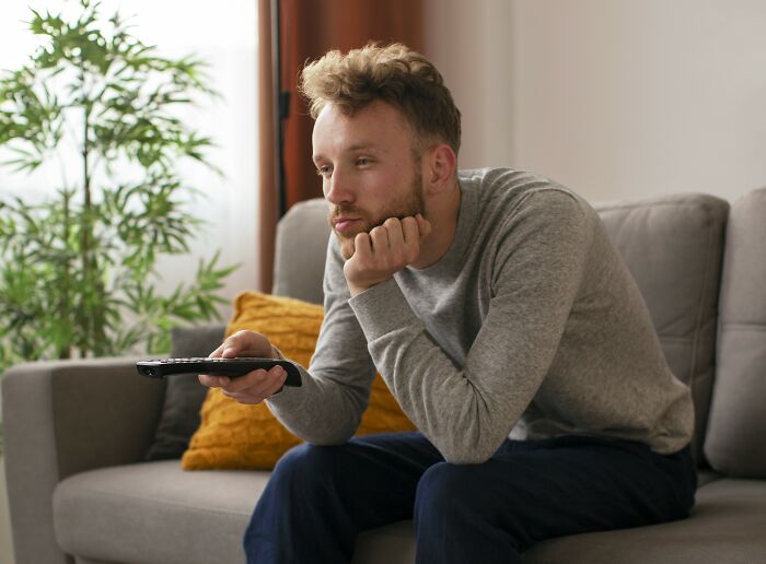 A person in a gray shirt sits on a couch, pondering unsolved invention ideas, holding a remote.