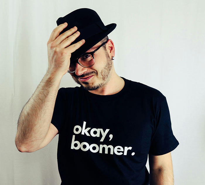 Hombre con gafas y barba vistiendo camiseta 'okay, boomer', tocando su sombrero negro, señal de humor tras traumas.