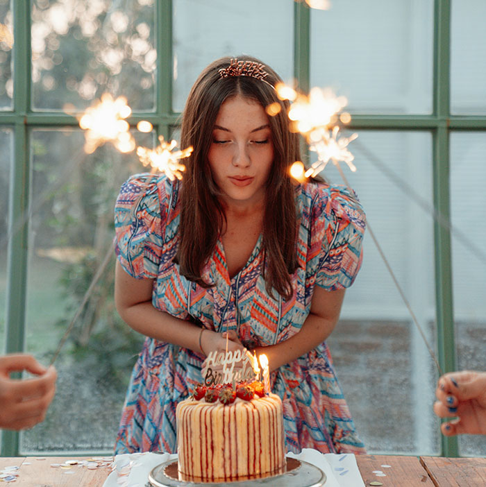 Mujer joven en cumpleaños, rodeada de bengalas, soplando velas de un pastel, celebrando momentos especiales.