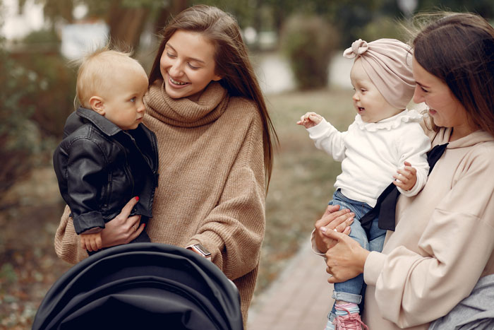 Women with babies in a park, engaging and smiling, highlighting feelings of being upstaged by a nephew's achievements.