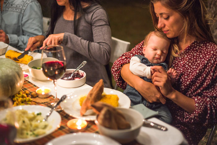 Woman holding baby at a dinner table, compares with premature nephew, surrounded by food and family.
