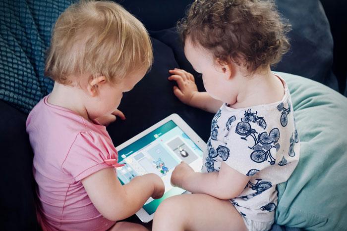 Two toddlers on a couch, interacting with a tablet, showcasing sibling dynamics and childhood development.