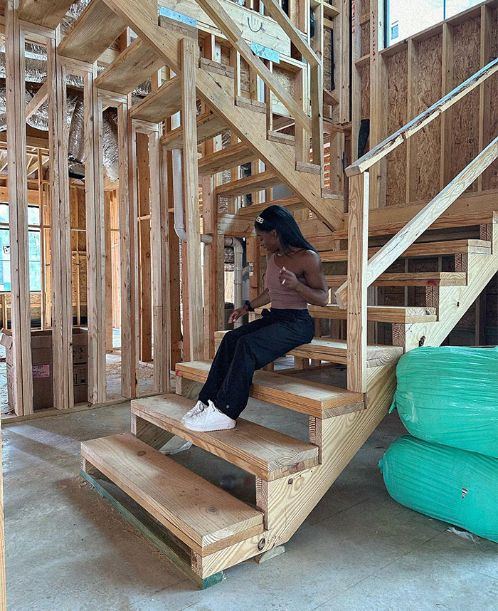 Simone Biles sitting on unfinished wooden stairs in a construction site, symbolizing building a fortune beyond the mat.