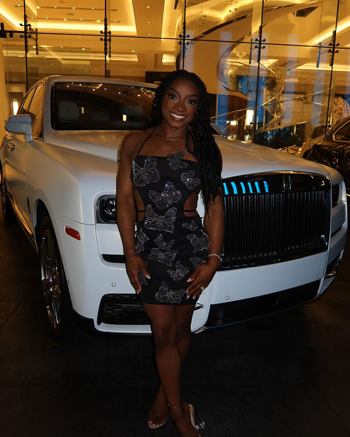 Olympic gymnast Simone Biles posing in front of a luxury car, wearing a black dress with butterfly patterns.