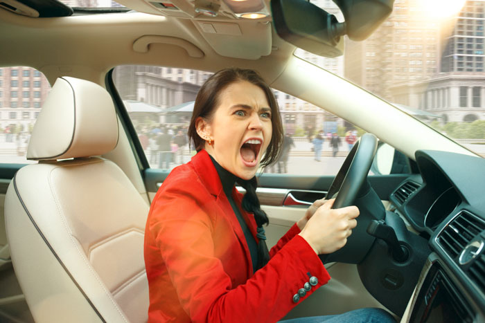 A woman in a red jacket behind the wheel, visibly upset, inside a car on a city street.