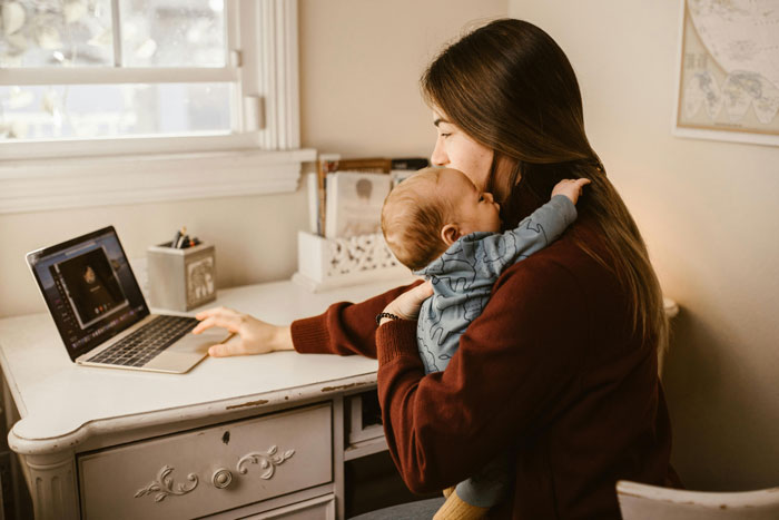 A woman holding a baby while using a laptop, representing the challenges of babysitting duties. A woman holding a baby while using a laptop, representing the challenges of babysitting duties.