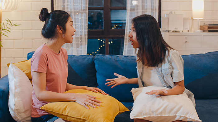 Two women on a sofa arguing, each holding a cushion, discussing boundaries and babysitting responsibilities.