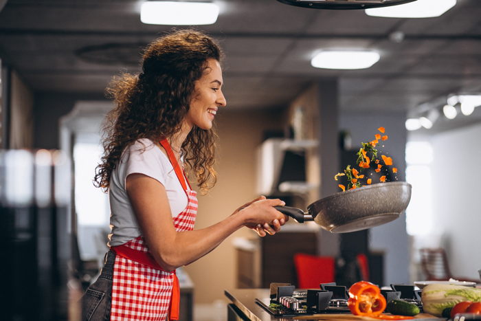 Woman cooking with skillet in modern kitchen, wearing a red apron, smiling while tossing vegetables in the air.