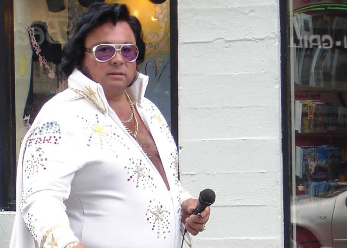 Elvis impersonator in a white jumpsuit, holding a microphone, standing outside a store, related to small town incident.