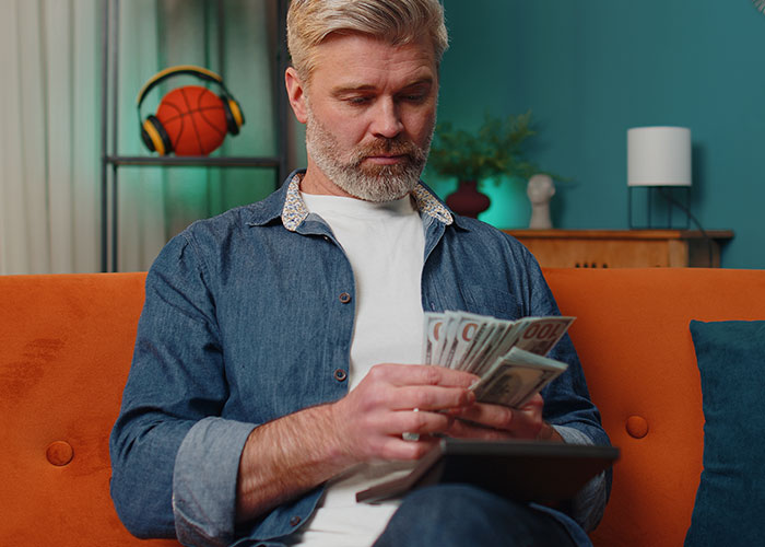 Man in denim shirt counting money, seated on orange couch, in a colorful living room.