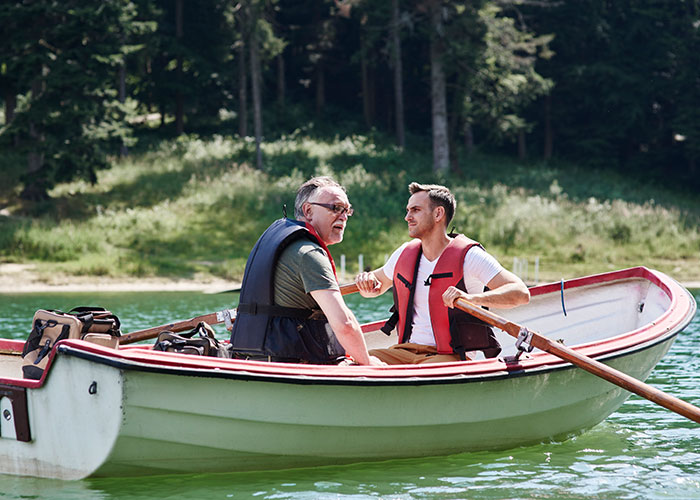 Two men in life jackets talking on a boat on a lake, surrounded by trees, possibly discussing a small town incident.