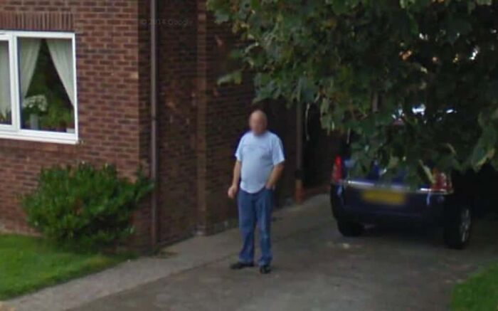 A person standing in a driveway captured by Google Earth near a brick house and parked car.