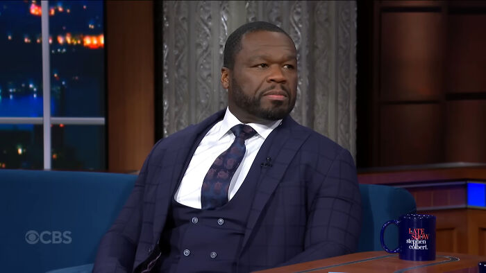 A celebrity in a dark suit on a talk show set, looking to the side, with a late-night show mug in view.
