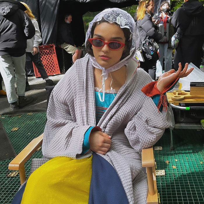 Actress on set in costume for 'Snow White,' wearing sunglasses and a bonnet, sitting casually in a director's chair.