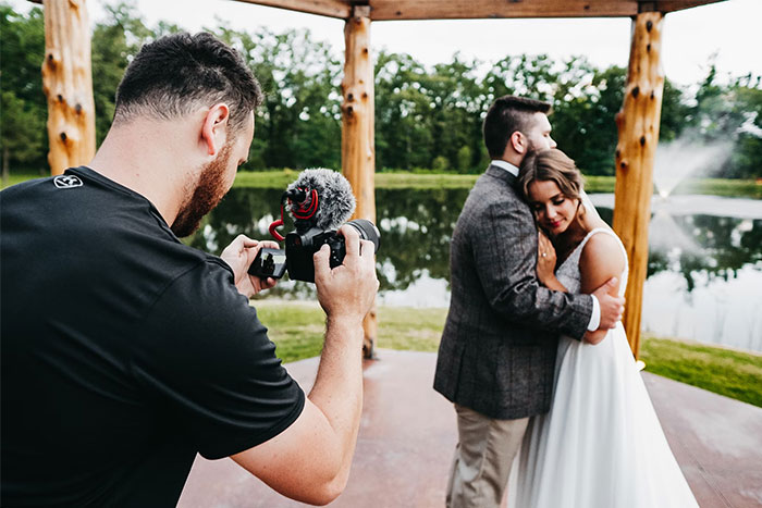 Wedding couple posing for a camera by a lake, with a videographer capturing the moment during a multi-day celebration. Wedding couple posing for a camera by a lake, with a videographer capturing the moment during a multi-day celebration.