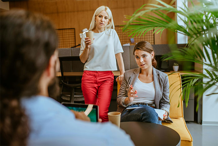Two women with coffee cups engaged in conversation at a modern indoor setting, featuring warm colors and greenery. Two women with coffee cups engaged in conversation at a modern indoor setting, featuring warm colors and greenery.