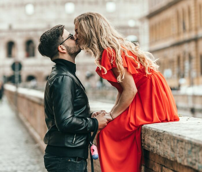 A couple kissing on a bridge, with the woman in a red dress and the man in a leather jacket.