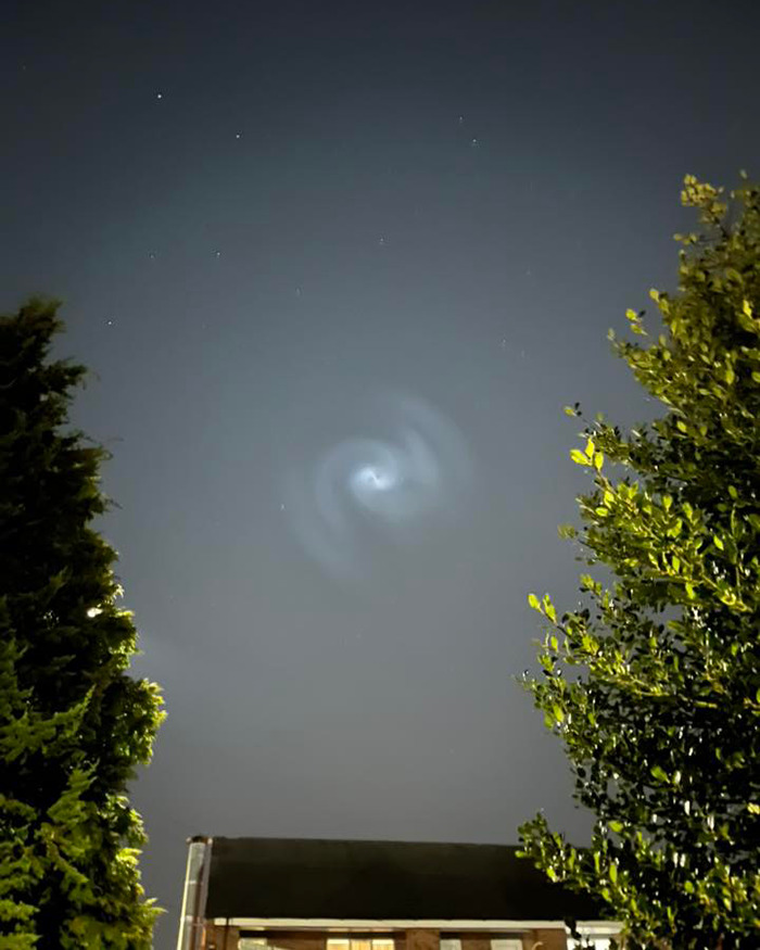 Mysterious spiral appears in night sky above UK, framed by trees and rooftops.