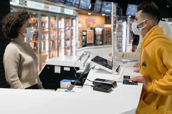 Moviegoer at a concession stand, wearing a mask and yellow hoodie, speaking to a staff member during a transaction.