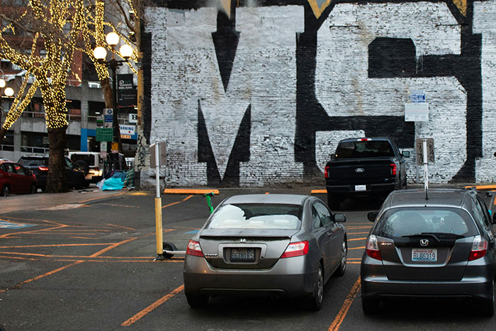 Coches estacionados en una calle urbana frente a un mural con letras grandes, personas que aún siguen vivas.