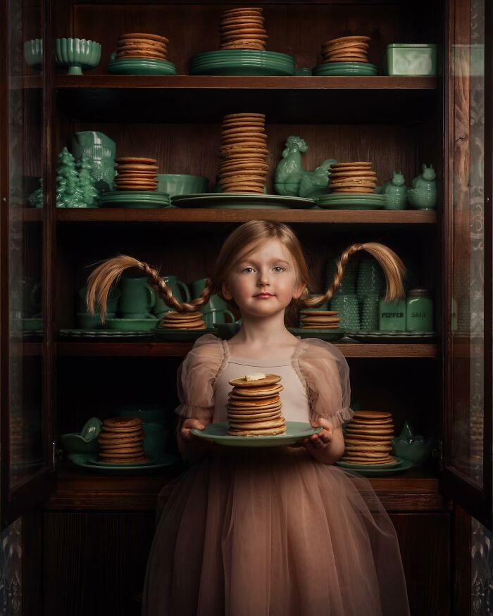Young girl holding pancakes, standing in front of a cabinet filled with green dishes, highlighting the magic of childhood.