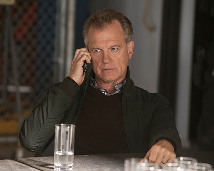 Disgraced actor on phone, seated at table, wearing a dark jacket and brown sweater.