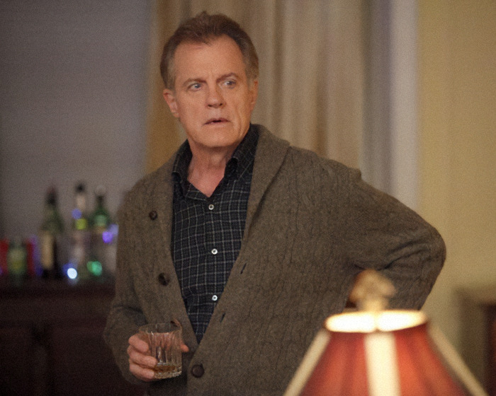 Disgraced actor in a sweater holding a drink, standing in a softly lit room with a lamp and bottles in the background.