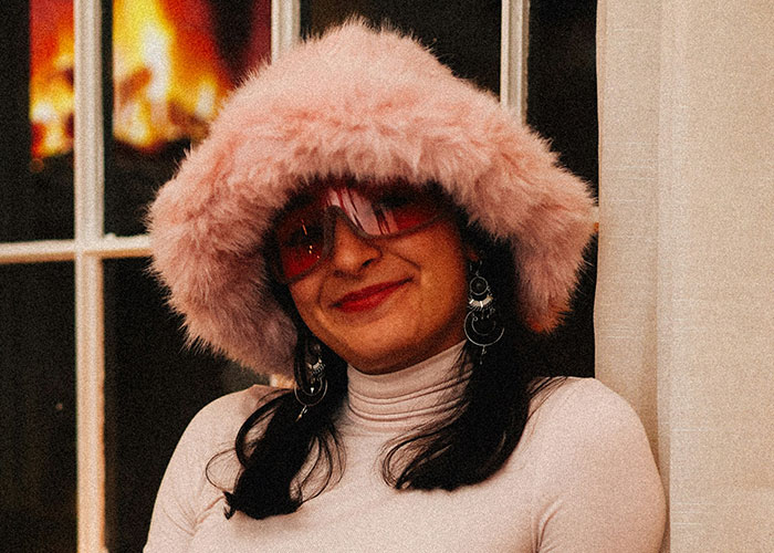 A woman wearing a pink furry hat and sunglasses indoors, smiling at the camera.