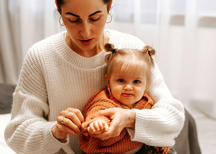 A woman in a white sweater with a baby in an orange sweater, indoors.