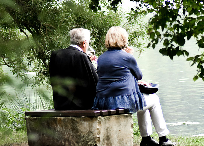 Elderly couple sit on a bench by a lake, surrounded by trees, illustrating peaceful moments.