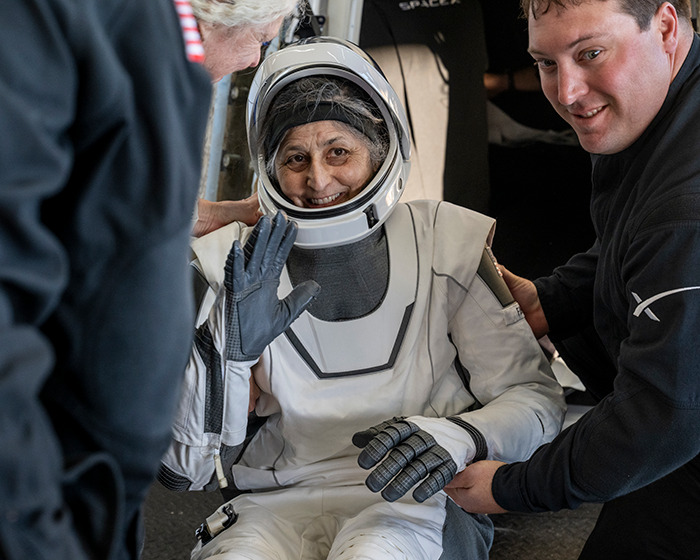 Astronaut appearing frail, waving in space suit, assisted by two people, raising doctors' health concerns.