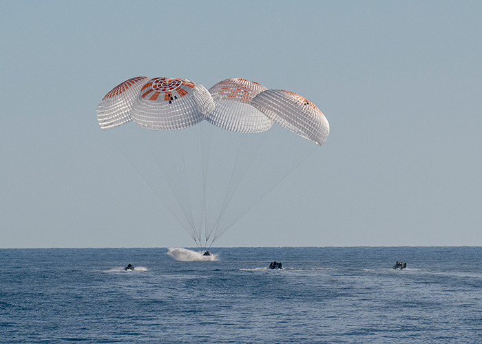 Parachutes slowing astronaut capsule as it lands in the ocean, with boats approaching for recovery.