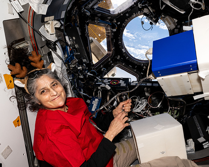 NASA astronaut in spacecraft, wearing festive antlers, with Earth visible through the window.
