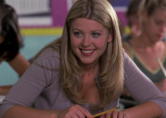 American Pie actress in a classroom setting, smiling and holding a pencil during a scene from the movie.