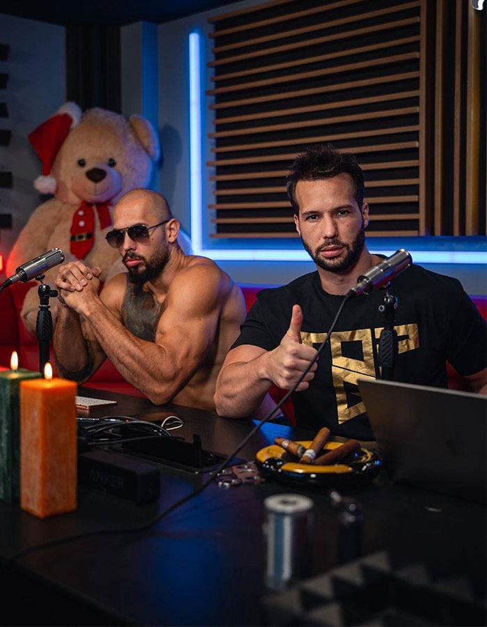 Two men podcasting at a table with microphones, candles, and a large teddy bear in the background. Two men podcasting at a table with microphones, candles, and a large teddy bear in the background.
